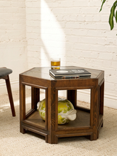 Load image into Gallery viewer, A hexagonal dark brown wooden end table with a glass top and open lower shelf, styled in a bright room with a white brick wall. A glass of beer, a book, and a green-and-white decorative object sit on and inside the table.
