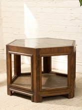 Load image into Gallery viewer, A hexagonal wooden end table with a dark brown finish, featuring a glass top and an open lower shelf, set against a white brick wall and beige carpet.
