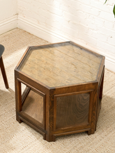 Load image into Gallery viewer, A hexagonal wooden side table with a clear glass top and lower shelf, set on a beige textured rug against a white brick wall.
