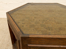 Load image into Gallery viewer, Close-up of an octagonal wooden table with a dark brown wood frame and a textured, patterned cork or jute top, set against a light beige wall and textured floor.
