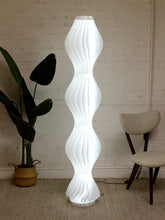 Load image into Gallery viewer, A modern white spiral floor lamp with a sculptural, wavy ribbed design stands in a room corner against a white brick wall, flanked by a potted plant on the left and a beige chair on the right.
