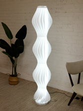 Load image into Gallery viewer, A tall, modern spiral floor lamp with a white ribbed shade, glowing with a warm light in a room corner with a brick wall, potted plant, and chair.

