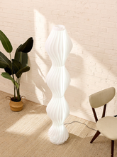 Load image into Gallery viewer, A modern white floor lamp with a sculptural, wavy ribbed design stands on a beige textured rug in a brightly lit room with a white brick wall. A potted green plant is on the left, and a cream-colored chair is on the right.
