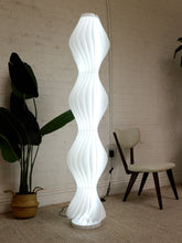 Load image into Gallery viewer, A tall, sculptural white floor lamp with a wavy, ribbed spiral design stands on a beige carpet in a room with a white brick wall, next to a potted plant and a wooden chair.
