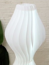 Load image into Gallery viewer, A white sculptural floor lamp with a fluted, pleated design, shown against a white brick wall background.
