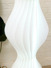 Load image into Gallery viewer, A close-up of a white modern floor lamp with a sculptural, ribbed, fluted design, set against a white brick wall background.
