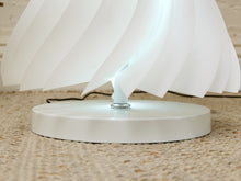 Load image into Gallery viewer, A close-up of a modern white table lamp with a circular base and layered, petal-like shade panels, shown on a woven beige rug.

