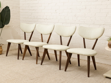 Load image into Gallery viewer, Five mid-century modern dining chairs with cream upholstered seats and curved backs on dark wood legs, arranged against a white brick wall.
