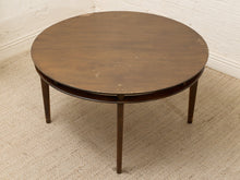 Load image into Gallery viewer, A round mid-century modern wooden coffee table with a dark brown finish, featuring a lower shelf and tapered legs, set on a textured beige rug against a white brick wall.
