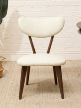Load image into Gallery viewer, A mid-century modern dining chair with a cream-colored textured fabric upholstery and dark brown wooden legs, set against a white brick wall and beige jute rug.
