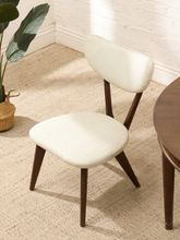 Load image into Gallery viewer, A mid-century modern dining chair with cream upholstered seat and backrest, supported by dark brown wooden legs, shown in a styled room setting on a beige rug next to a wooden table.
