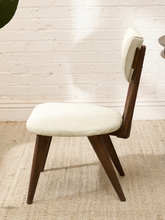Load image into Gallery viewer, A mid-century modern dining chair with a dark brown wooden frame and cream-colored upholstered seat and backrest, set on a woven jute rug against a white brick wall.
