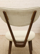 Load image into Gallery viewer, A mid-century modern wooden dining chair viewed from the back, featuring a curved upholstered backrest and seat cushion in a textured cream fabric, supported by a dark brown wooden frame against a white brick wall.
