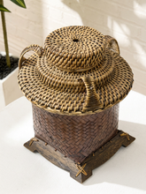 Load image into Gallery viewer, A woven natural-fiber storage basket with a matching lid, shown on a white surface against a white brick wall. The basket has a dark brown base and sits on a wooden stand.
