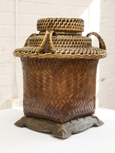 Load image into Gallery viewer, A traditional hand-woven bamboo basket with a lid, side handles, and a dark wooden base, photographed against a white brick wall background.
