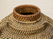 Load image into Gallery viewer, A close-up of a hand-woven natural fiber basket with a circular woven rim and a light tan horizontal accent band, set against a light off-white brick wall background.
