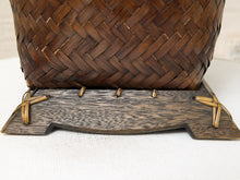 Load image into Gallery viewer, A close-up of a dark brown woven basket with a decorative wooden base featuring stitched details and tie accents, set on a white textured fabric surface.
