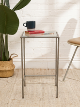 Load image into Gallery viewer, A square metal side table with a clear glass top, styled with a navy and white mug, a red book, and a pen. The table is set on a beige woven rug against a white brick wall, with a potted plant on the left and a chair on the right.
