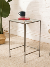 Load image into Gallery viewer, A square side table with a clear glass top and a slim silver metal frame, styled in a room setting with a white brick wall, beige rug, a potted plant, and a mug on top.

