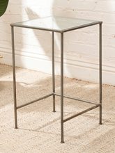 Load image into Gallery viewer, A square side table with a clear glass top and a weathered gray metal frame, set on a beige woven rug against a white brick wall.
