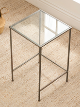 Load image into Gallery viewer, A square side table with a clear glass top and a slim silver metal frame, shown on a beige woven rug in a room setting.
