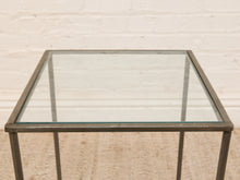 Load image into Gallery viewer, A square metal-framed glass table with a dark gray or black frame and a clear glass top, shown against a light beige brick wall background.
