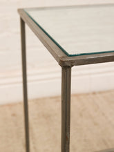 Load image into Gallery viewer, Close-up of a rectangular table corner with a clear glass top and a distressed gray metal frame, set against a light beige wall and floor.
