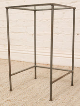 Load image into Gallery viewer, A square metal side table with a clear glass top and a rustic dark gray painted frame, set against a white brick wall and light woven rug.
