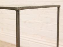Load image into Gallery viewer, Close-up of a rustic industrial table frame with a glass top, featuring weathered dark gray metal legs and joints against a white brick wall background.
