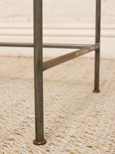 Load image into Gallery viewer, Close-up of a rustic metal table leg with a weathered, rusted finish, standing on a textured beige woven rug against a white brick wall.
