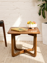 Load image into Gallery viewer, A mid-century modern wooden side table with a warm medium-brown finish, styled with a book and a white bowl of fruit on a textured beige rug against a white brick wall.
