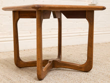Load image into Gallery viewer, A mid-century modern wooden side table with a warm medium-brown finish, featuring a rounded rectangular top and curved legs, set on a textured beige rug against a white brick wall.
