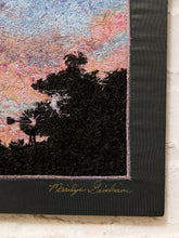Load image into Gallery viewer, A close-up of a textile wall hanging or tapestry showing a dark tree silhouette against a pastel sky with pink, blue, and lavender tones. The piece has a wide black border with a gold signature reading 'Nadine Graham' near the bottom, displayed against a light brick wall.
