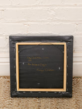 Load image into Gallery viewer, The back of a framed artwork, showing a black fabric-covered backing with handwritten yellow text and a light wood frame, leaning against a white brick wall on a woven rug.
