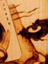 Load image into Gallery viewer, Close-up of a wooden card featuring the word 'JOKER' vertically on the left, with a stylized face and dark eye graphic burned or printed onto the wood in warm brown tones.
