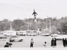 Load image into Gallery viewer, Black and white photograph of a jet airplane flying above a street with buses, cars, utility poles, and several small figures of people in the foreground.
