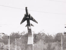 Load image into Gallery viewer, Black and white photograph of a jet airplane mounted on a tall pole as a monument, with trees and utility wires visible in the background.
