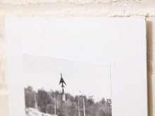 Load image into Gallery viewer, A black and white photographic print mounted on a large white backing board, leaning against a textured light beige brick wall.
