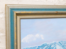 Load image into Gallery viewer, A close-up of a framed landscape painting showing blue mountains and a pale sky, with a distressed blue and beige frame mounted on a textured white wall.
