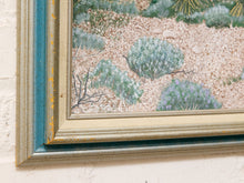 Load image into Gallery viewer, Close-up of a framed desert landscape painting showing green shrubs and plants over a sandy beige ground, with a distressed blue and gold frame against a white wall.
