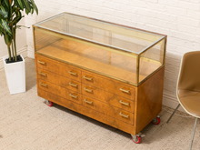 Load image into Gallery viewer, A mid-century modern wooden display cabinet with a glass top case, six drawers, brass handles, and red caster wheels, shown in a room setting next to a potted plant and a chair.
