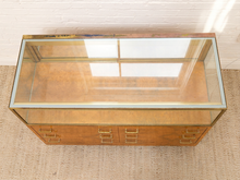 Load image into Gallery viewer, A mid-century modern glass display case with a warm wood-grain cabinet base, brass hardware, and a clear glass top, shown on a textured beige rug against a white brick wall.
