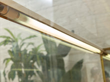 Load image into Gallery viewer, A close-up of a long, slim LED strip light glowing with a warm white light, mounted on a metallic gold frame above a glass surface with reflections of indoor plants.
