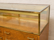 Load image into Gallery viewer, A vintage wooden display case with a glass top, brass frame, and drawers below, set against a white brick wall.
