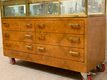 Load image into Gallery viewer, A vintage wooden rolling cabinet with six drawers, brass handles, and a glass display top, shown on casters in an indoor setting.
