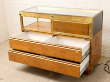 Load image into Gallery viewer, A vintage mid-century wooden display cabinet with a glass top, brass frame, and three open drawers showing white interiors, set against a white brick wall and beige carpet.
