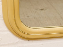 Load image into Gallery viewer, A close-up of a vintage-style wall mirror with a glossy mustard yellow frame, resting against a white painted brick wall.
