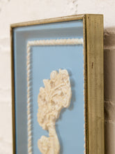 Load image into Gallery viewer, A close-up of a framed decorative wall art piece with a distressed gold frame, light blue backing, white rope border, and a cream-colored sculpted floral motif, shown against a white brick wall.

