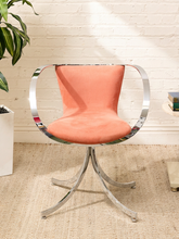 Load image into Gallery viewer, A modern swivel chair with a peach-pink upholstered seat and curved backrest, supported by a polished chrome metal base and integrated armrests, set against a white brick wall on a beige rug.
