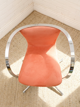 Load image into Gallery viewer, A mid-century modern accent chair with a curved, polished chrome tubular frame and a coral pink upholstered seat, shown on a textured beige rug in the corner of a room with white paneled walls.
